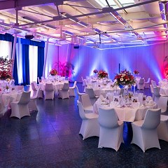 Kleiner Saal mit Eventbeleuchtung und festlich gedeckten runden Tische und Stühle mit weißen Hussen für eine private Feier