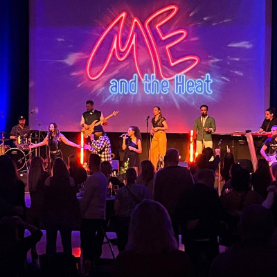 9-köpfige Band rockt in abgedunkeltem Raum die Bühne vor jubelndem und tanzendem Publikum, dahinter ist groß der Schriftzug des Logos in den Leuchtfarben pink, blau und lila eingeblendet