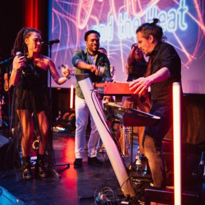Zu sehen ist der Keyboarder mit Sängerin Christine und einem Gastsänger auf der Bühne zu sehen, dahinter ist der Schriftzug des Bandlogos in den Leuchtfarben pink, weiß, blau, lila zu sehenl